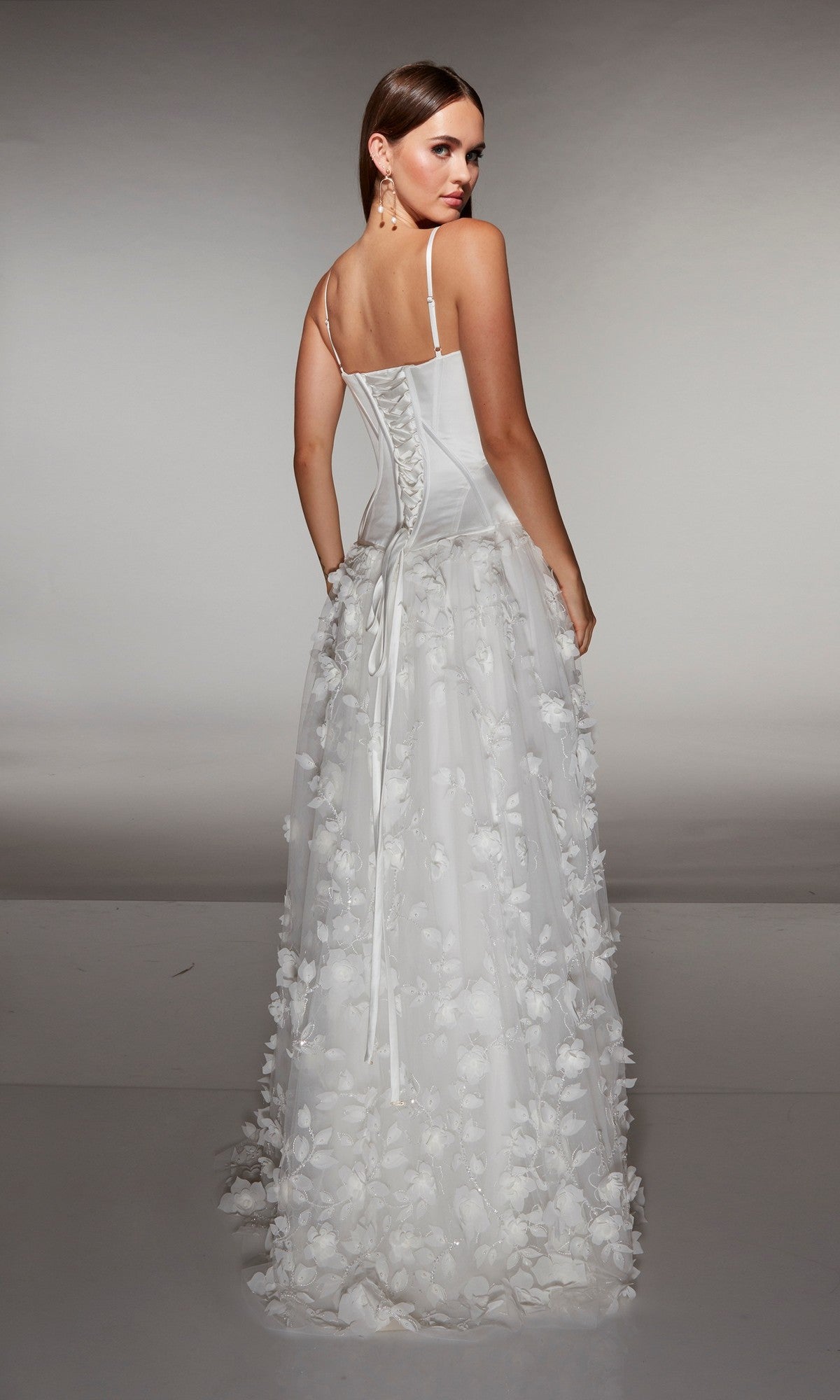 A woman stands facing away, wearing the Alyce 62072 long formal dress—a white sleeveless gown with a corset bodice and 3D floral-lace appliques.